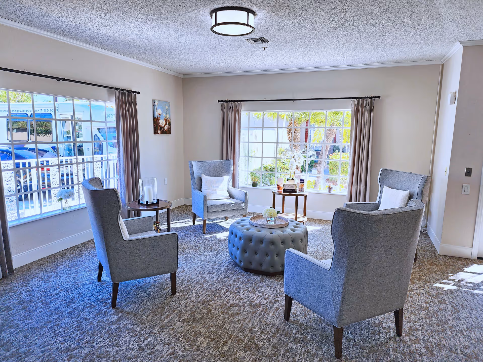 A bright and cozy sitting area with four gray upholstered armchairs arranged around a round tufted ottoman. The room has large windows with beige curtains letting in natural light, a small round side table with candles, and a small table with decorative items near the window. The carpet is patterned in neutral tones, and the walls are painted light beige.