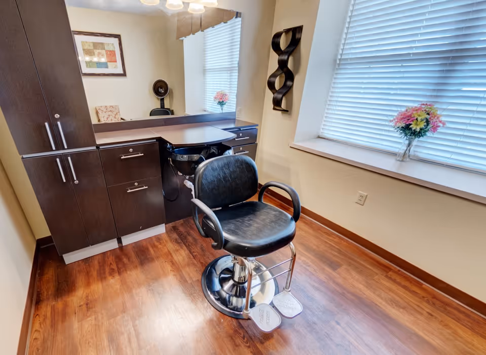 A hair salon station featuring a black styling chair in front of cabinets, a mirror, and a window with a vase of flowers.