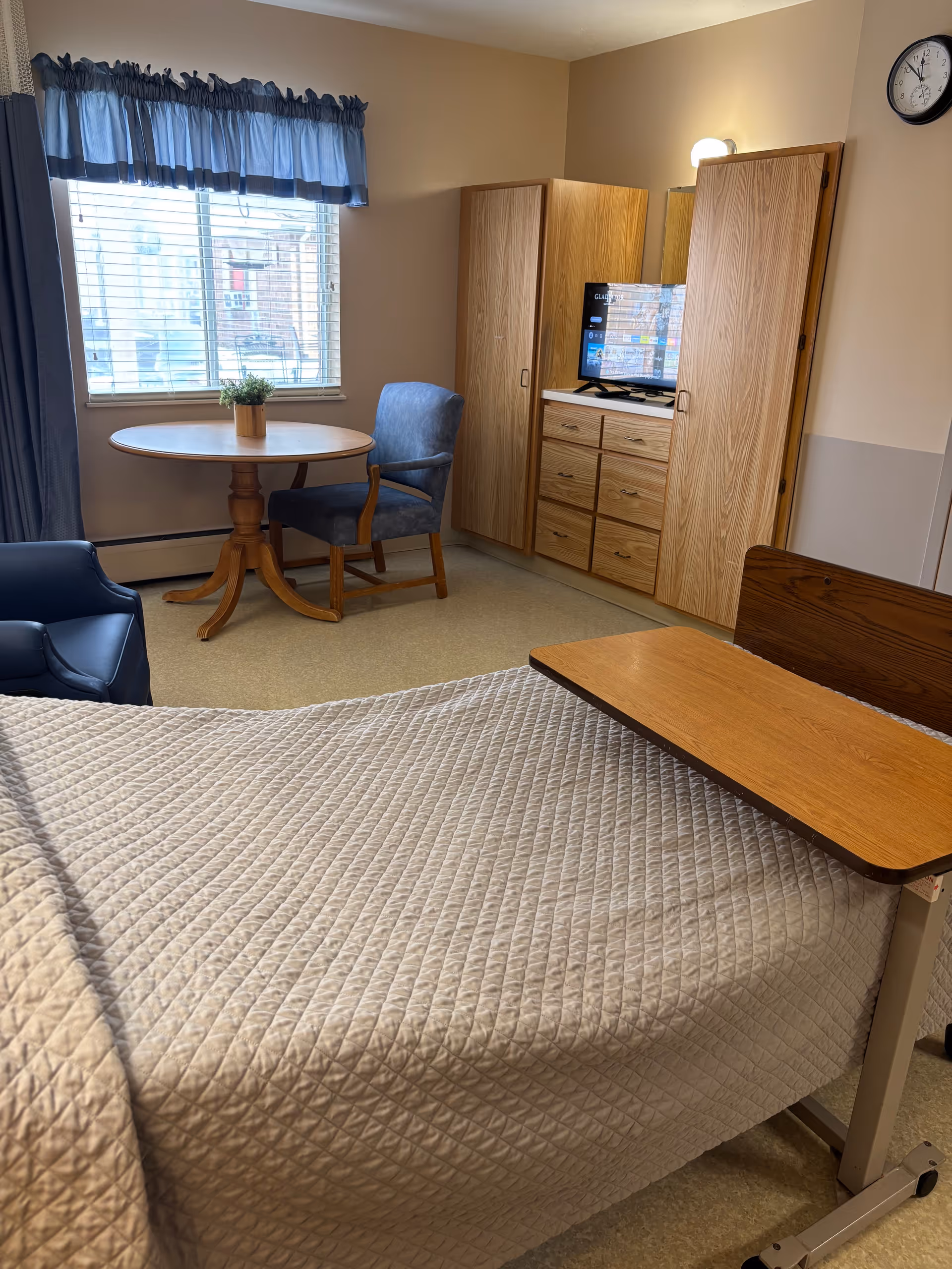 A senior living facility bedroom with a bed covered in a beige quilted blanket and an overbed table. There is a wooden round table with a small plant on it and a blue upholstered chair next to a window with blue curtains. Across from the bed is a wooden dresser with a TV on top, flanked by two wooden wardrobes. A clock is mounted on the wall above the dresser.