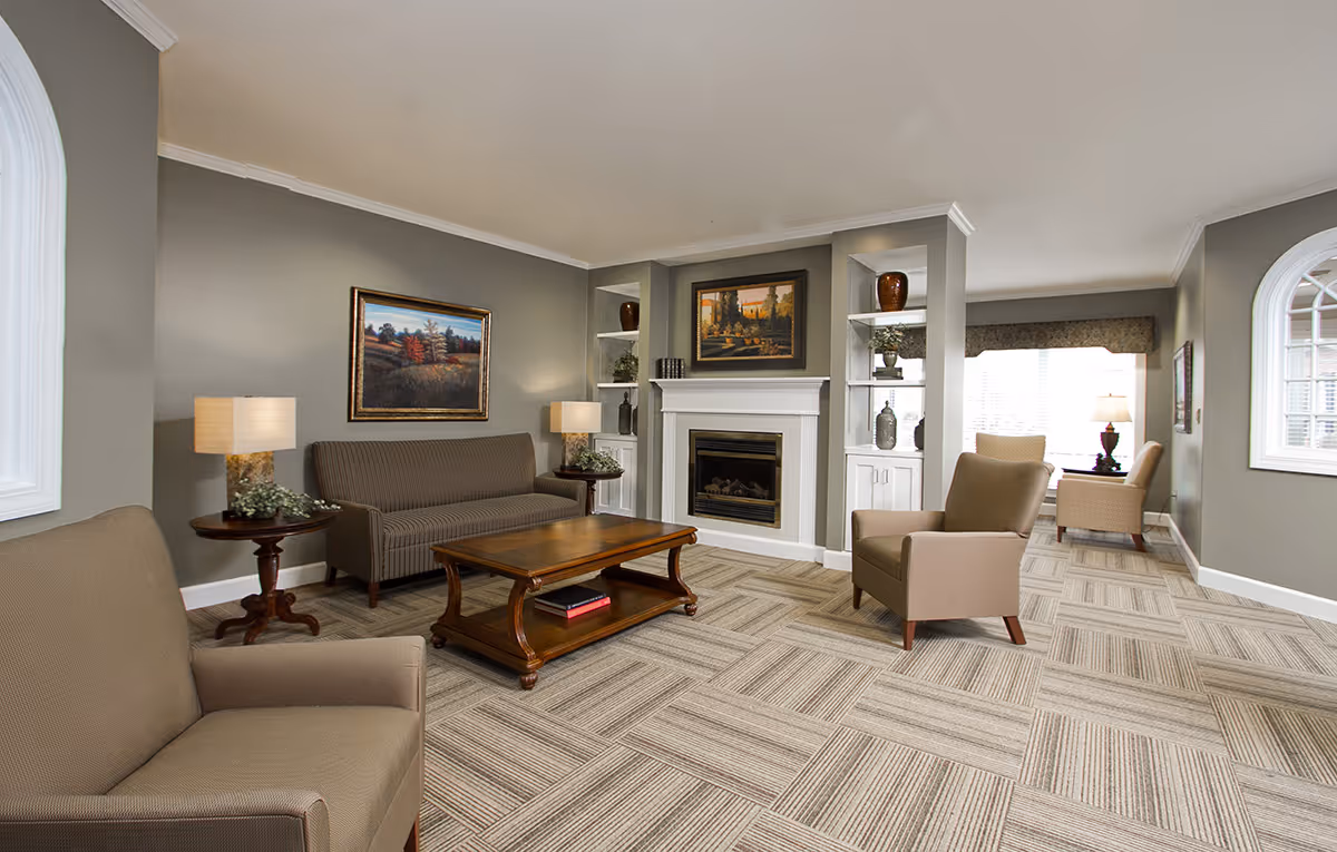 A cozy living room with beige armchairs and a striped sofa arranged around a wooden coffee table. The room features a fireplace with built-in shelves on either side, decorated with vases and plants. Two framed landscape paintings hang on the walls, and the room is well-lit with natural light coming through large windows.