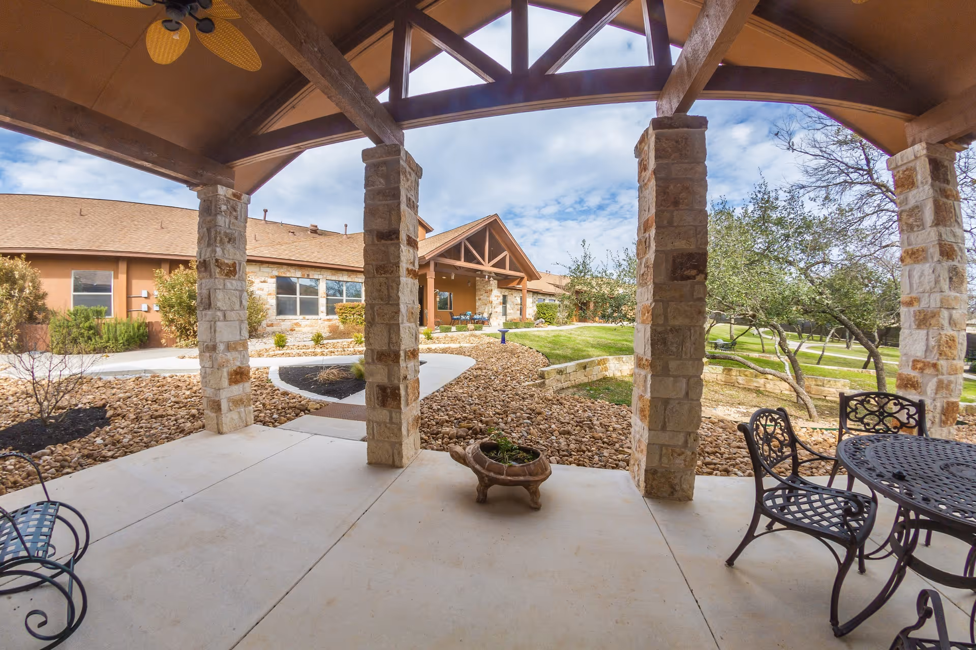 Covered stone-column patio with metal outdoor furniture looking toward the facility entrance and landscaped grounds.