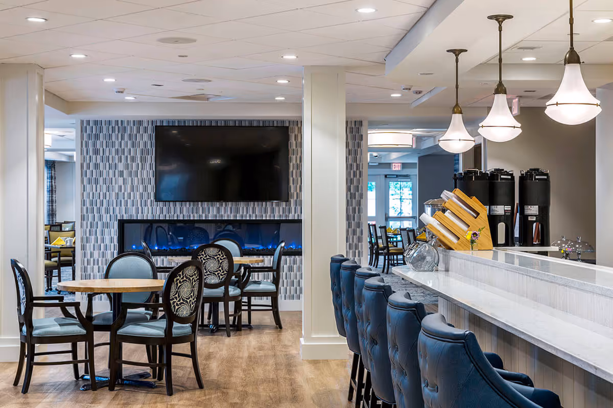 A modern common area with a bar counter featuring blue cushioned bar stools and pendant lights above. There are round tables with chairs, a wall-mounted flat screen TV, and a decorative electric fireplace beneath it. The space has a light wood floor and neutral-colored walls with patterned wallpaper behind the TV.