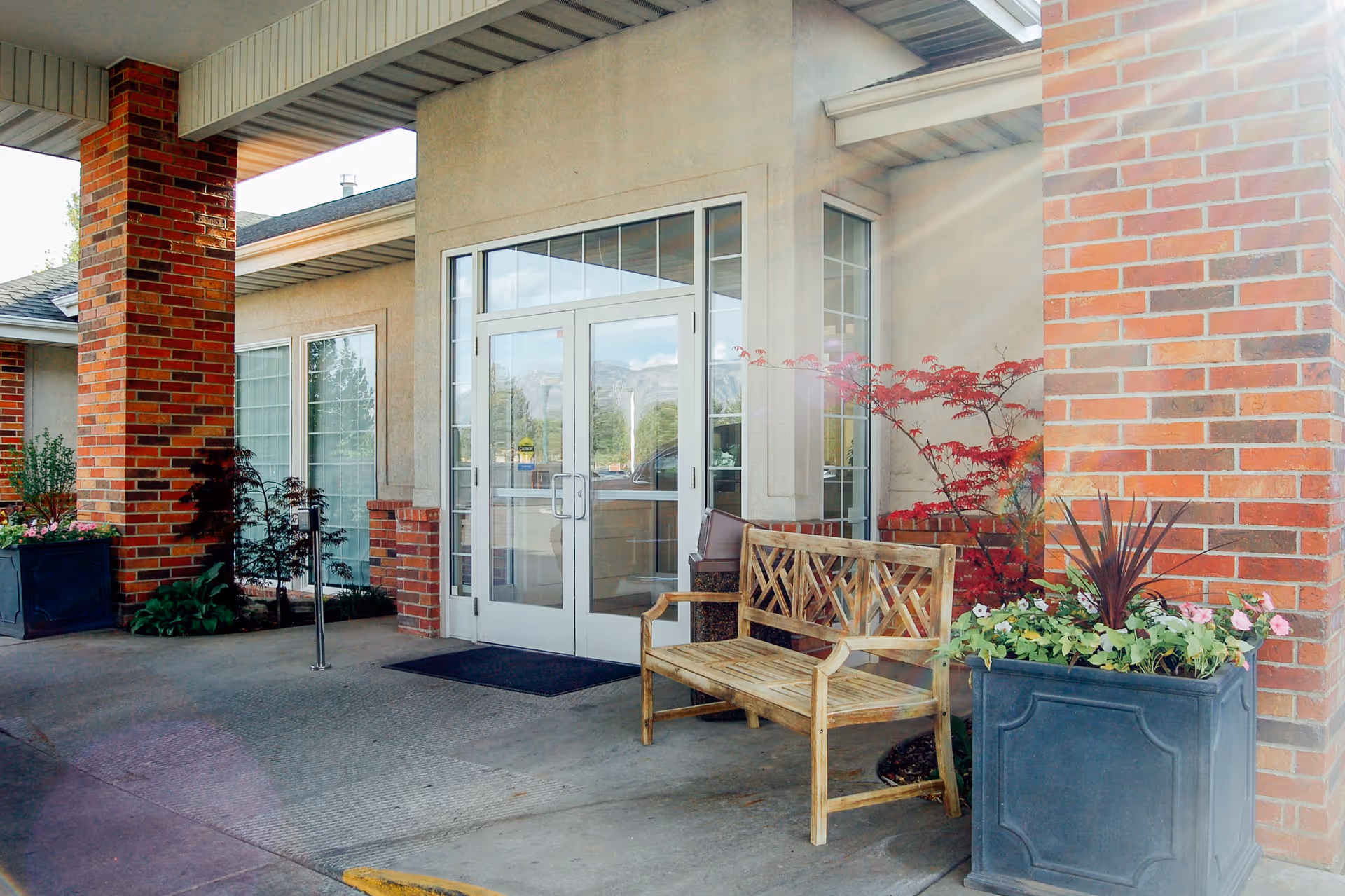 Entrance area of a building with double glass doors, brick pillars, a wooden bench, and large planters with flowers and plants on either side.