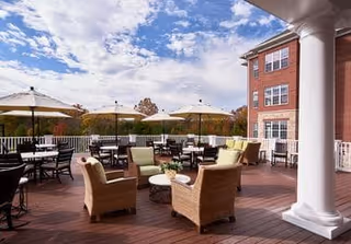 Outdoor patio area with wicker chairs and tables under large umbrellas on a wooden deck, adjacent to a multi-story brick building under a partly cloudy sky.