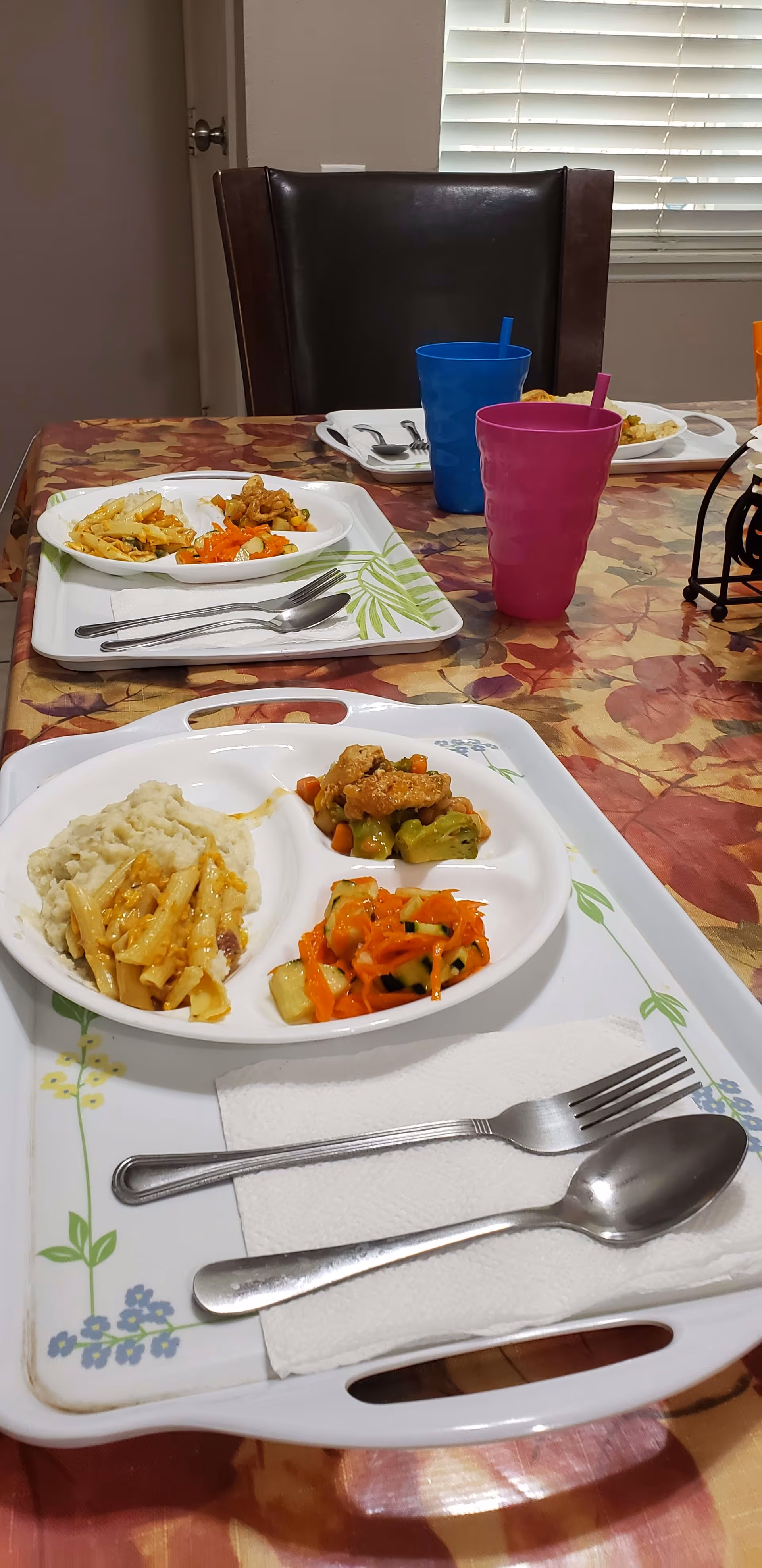 A dining table with a floral tablecloth set with trays of food. Each tray has a divided plate containing mashed potatoes, pasta, mixed vegetables, and a salad. There are utensils placed on napkins on the trays and colorful plastic cups with straws. A dark brown chair and a window with blinds are visible in the background.