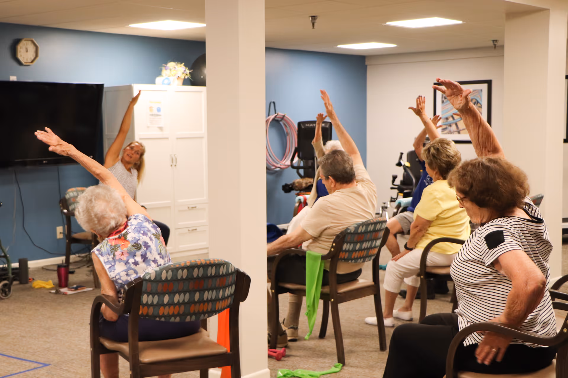 A group of elderly individuals seated on chairs in a room participating in a seated exercise class, stretching their arms overhead. The room has blue walls, a clock, a television, and exercise equipment in the background.