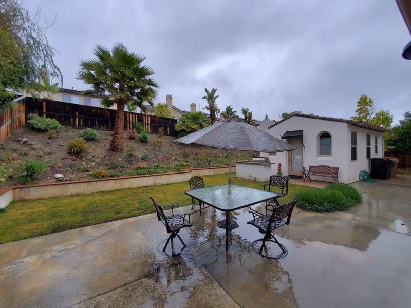 Outdoor patio area with a glass-top table and four metal chairs arranged around it. A large umbrella is centered over the table. The patio is wet from rain, and there is a small white building with a bench and some greenery nearby. In the background, there is a sloped garden with various plants and palm trees, enclosed by a wooden fence under a cloudy sky.