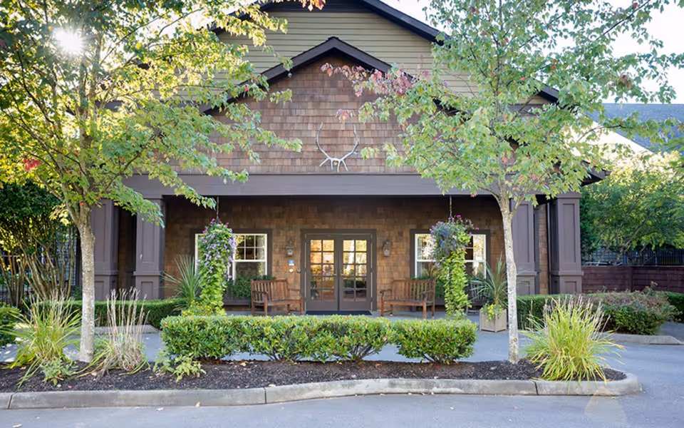 Front exterior view of Weatherly Inn Lake Meridian showing a building entrance with double doors, two wooden benches on the porch, hanging flower baskets, and landscaping including trees and shrubs.