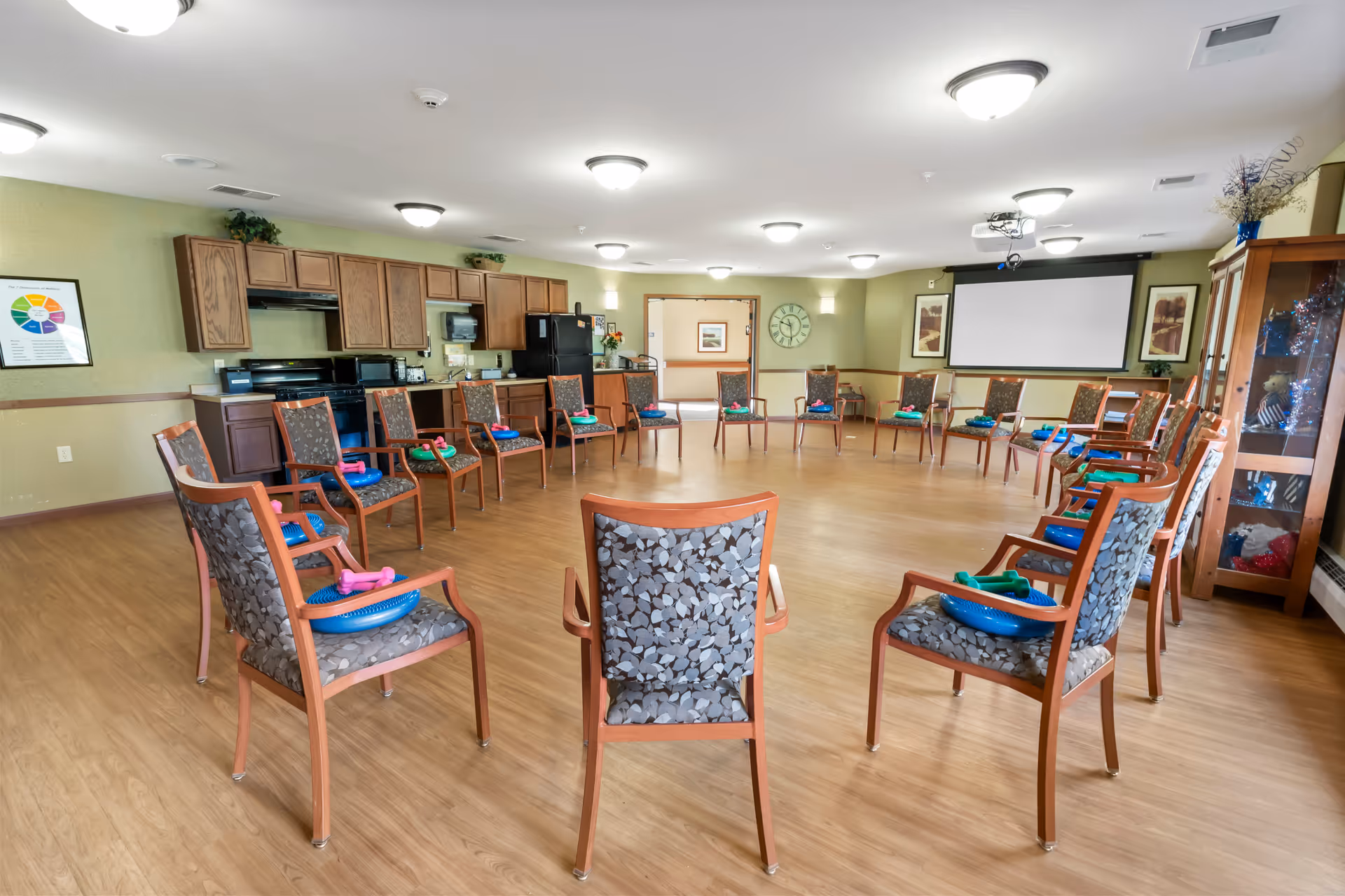 A spacious activity room in a senior living facility with chairs arranged in a large circle. Each chair has a blue plastic bowl and colorful exercise equipment placed on the seat. The room has wooden flooring, a kitchenette with cabinets, a refrigerator, and a stove along one wall. There is a large clock on the far wall, framed artwork, and a projector screen. The room is well-lit with ceiling lights and has a glass display cabinet on the right side.