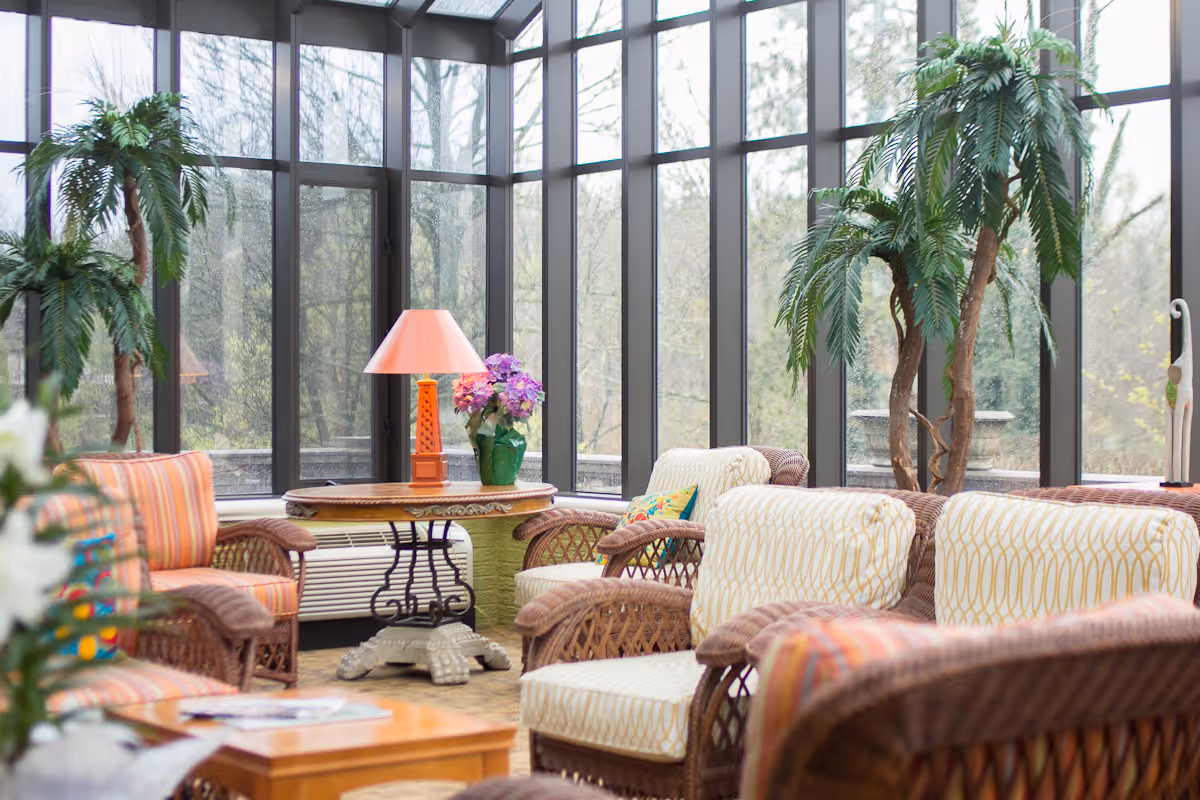 A bright sunroom with large floor-to-ceiling windows showing an outdoor view of trees. The room is furnished with wicker chairs and sofas with patterned cushions, a round wooden table with a pink lamp and a vase of flowers, and several potted plants.