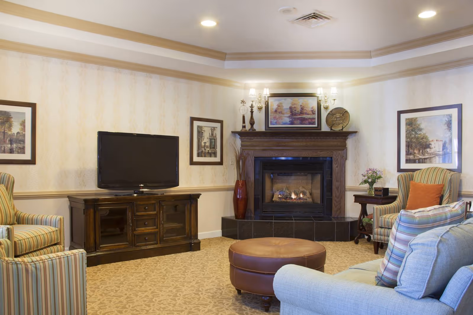 Cozy living room with a fireplace, flat-screen TV on a wooden console, striped armchairs, a round leather ottoman and framed artwork.