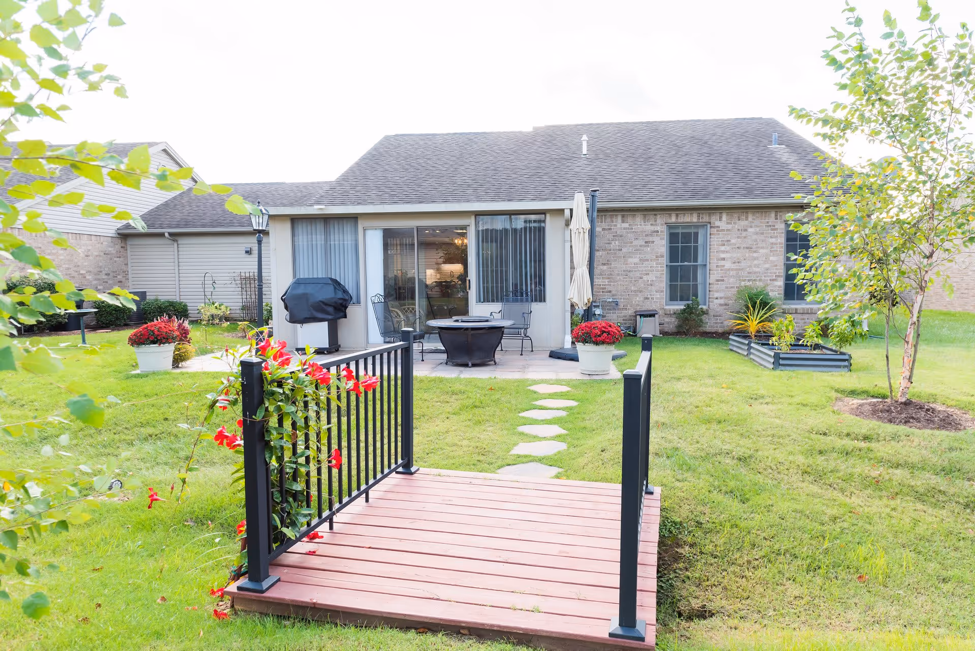 A backyard view of a single-story brick house with a small wooden bridge decorated with red flowers leading to a stone pathway. The patio area has outdoor furniture including chairs, a table, a grill, and a closed umbrella. The yard is green with grass, potted plants, and a small tree on the right side.