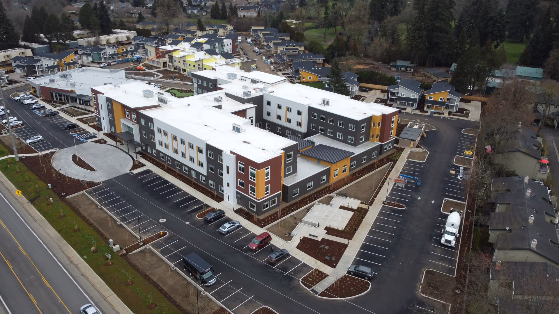 Aerial view of Marquis Eugene Assisted Living facility showing a large, modern multi-story building with white and colored exterior walls, surrounded by parking lots with several parked cars. The facility is located near a road and residential houses, with landscaped areas around the building.
