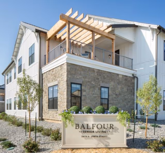 Exterior view of Balfour Central Park senior living facility showing a two-story building with white siding and stone accents, a balcony with wooden pergola, small trees, and a sign in front that reads 'BALFOUR SENIOR LIVING 2879 N. UINTA STREET'.