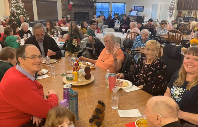 Residents and visitors seated around large tables in a decorated communal dining room enjoying a meal.