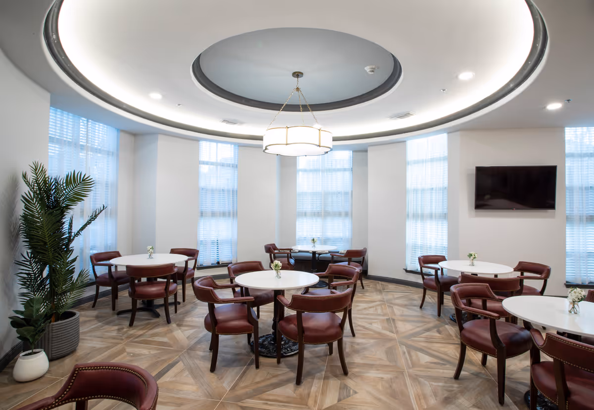 A bright, modern dining area with round white tables and maroon upholstered chairs arranged neatly. The room features large windows with sheer white curtains, a circular recessed ceiling with a central chandelier, wood-patterned flooring, a flat-screen TV mounted on the wall, and potted plants in the corner.
