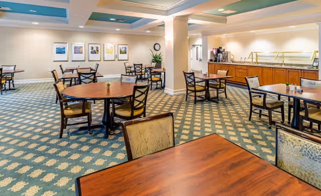 Spacious senior living dining room with round and rectangular tables, chairs, patterned carpet, and a buffet counter along the wall.