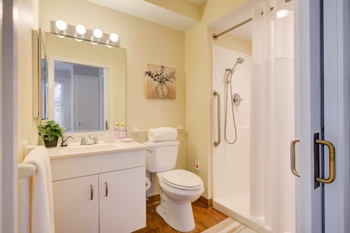 A clean and well-lit bathroom featuring a white vanity with a sink, a large mirror with lights above it, a toilet with a towel on the grab bar, and a walk-in shower with a handheld showerhead and a white curtain. The walls are painted light yellow and the floor has wood-like flooring.