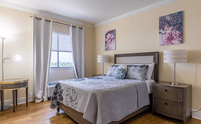 Bright furnished bedroom featuring a bed with decorative pillows, two nightstands with lamps, floral wall art, and a window with curtains.