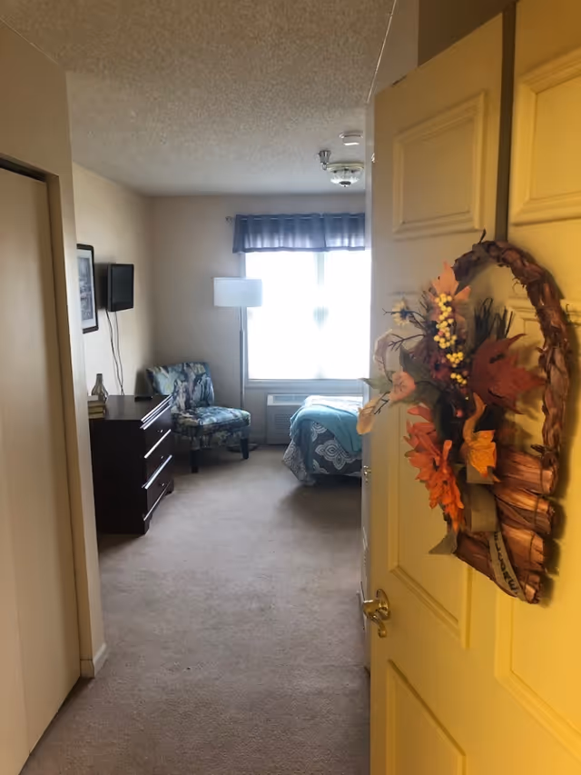 View through an open front door into a furnished bedroom with a bed, armchair, dresser, wall-mounted TV, and a fall wreath on the door.