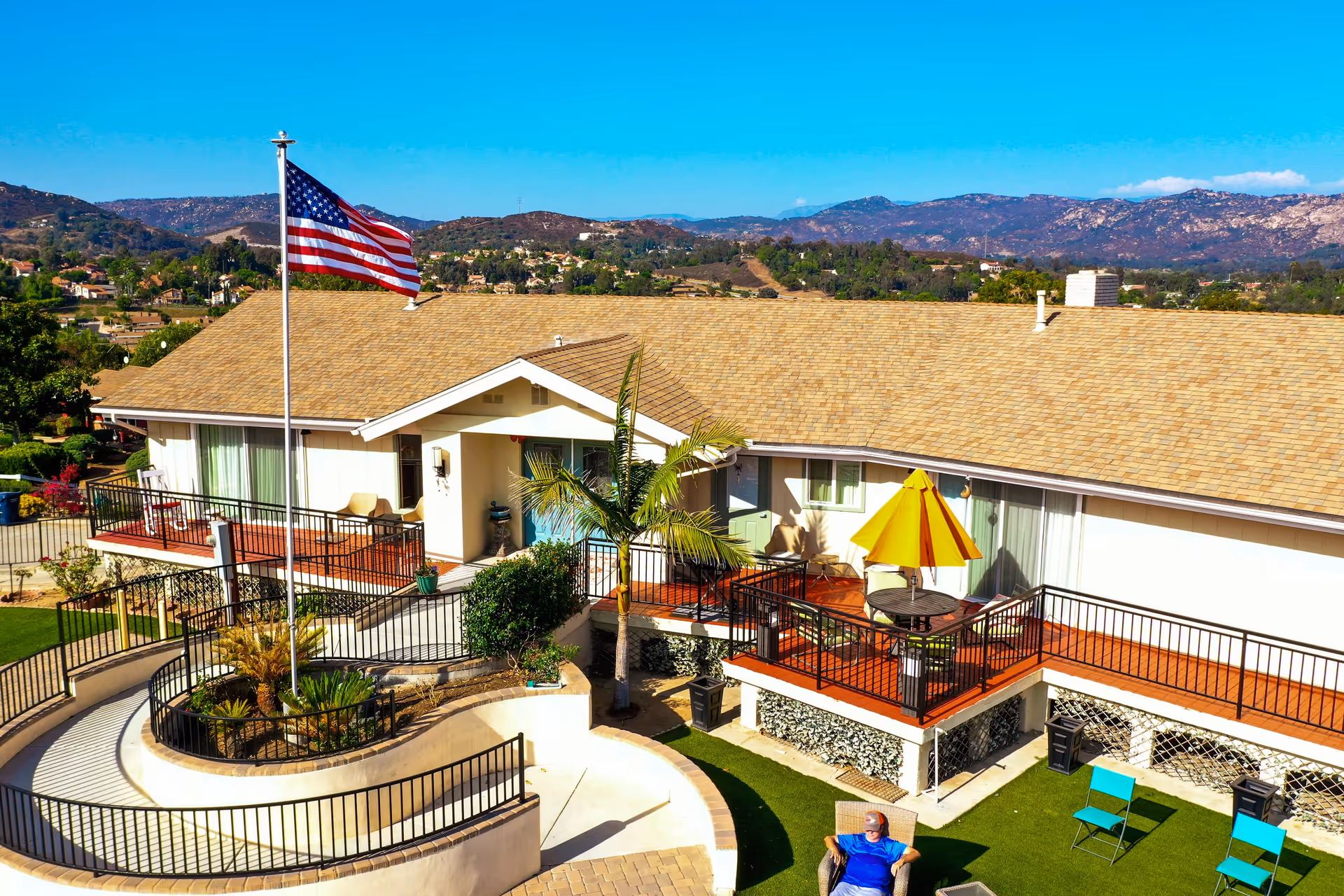 Outdoor view of Champine Manor RCFE showing a single-story building with a tan roof and white walls. There is a large American flag on a flagpole in front of the building. The area includes a raised wooden deck with chairs and a table with a yellow umbrella. A man is sitting on a chair on the green lawn below the deck. Surrounding the facility are hills and a clear blue sky.