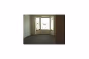 An empty room with a large window featuring three sections, allowing natural light to enter. The walls are plain and light-colored, and the floor appears to be carpeted.