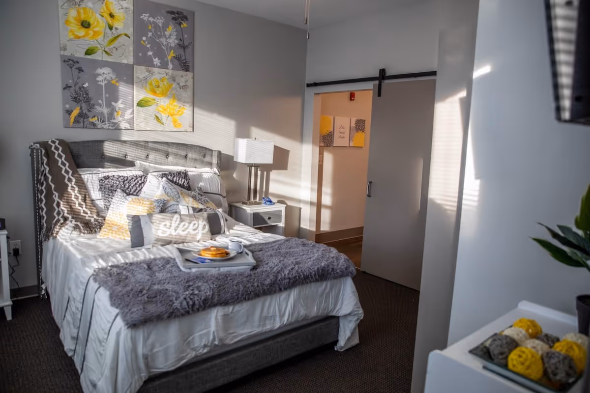 Sunlit furnished bedroom with a made bed, decorative pillows and artwork, a nightstand with a lamp, and a sliding barn door.