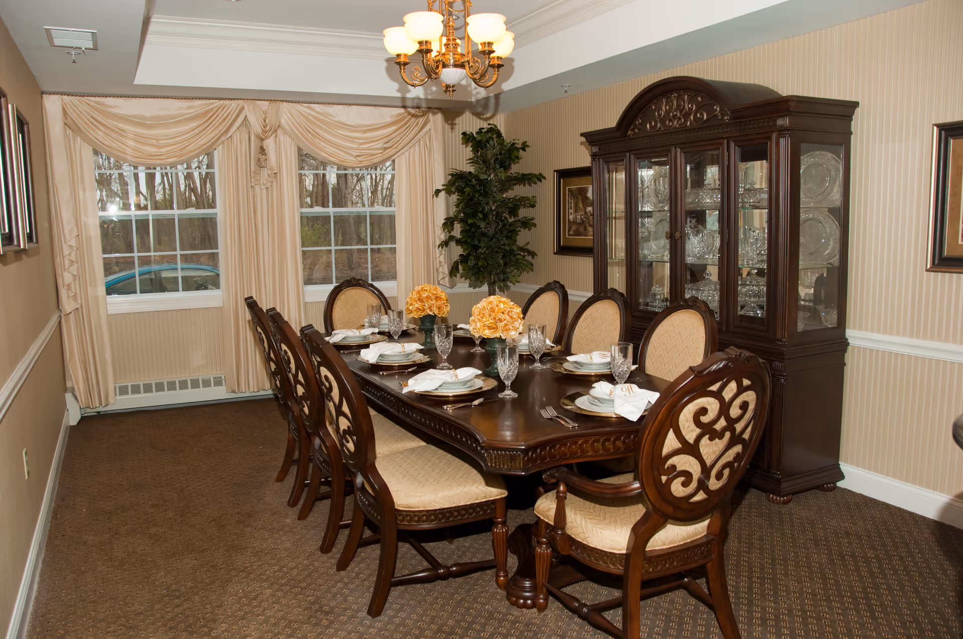 Formal dining room with a long wooden table set for a meal, ornate chairs, a china cabinet, and draped windows.