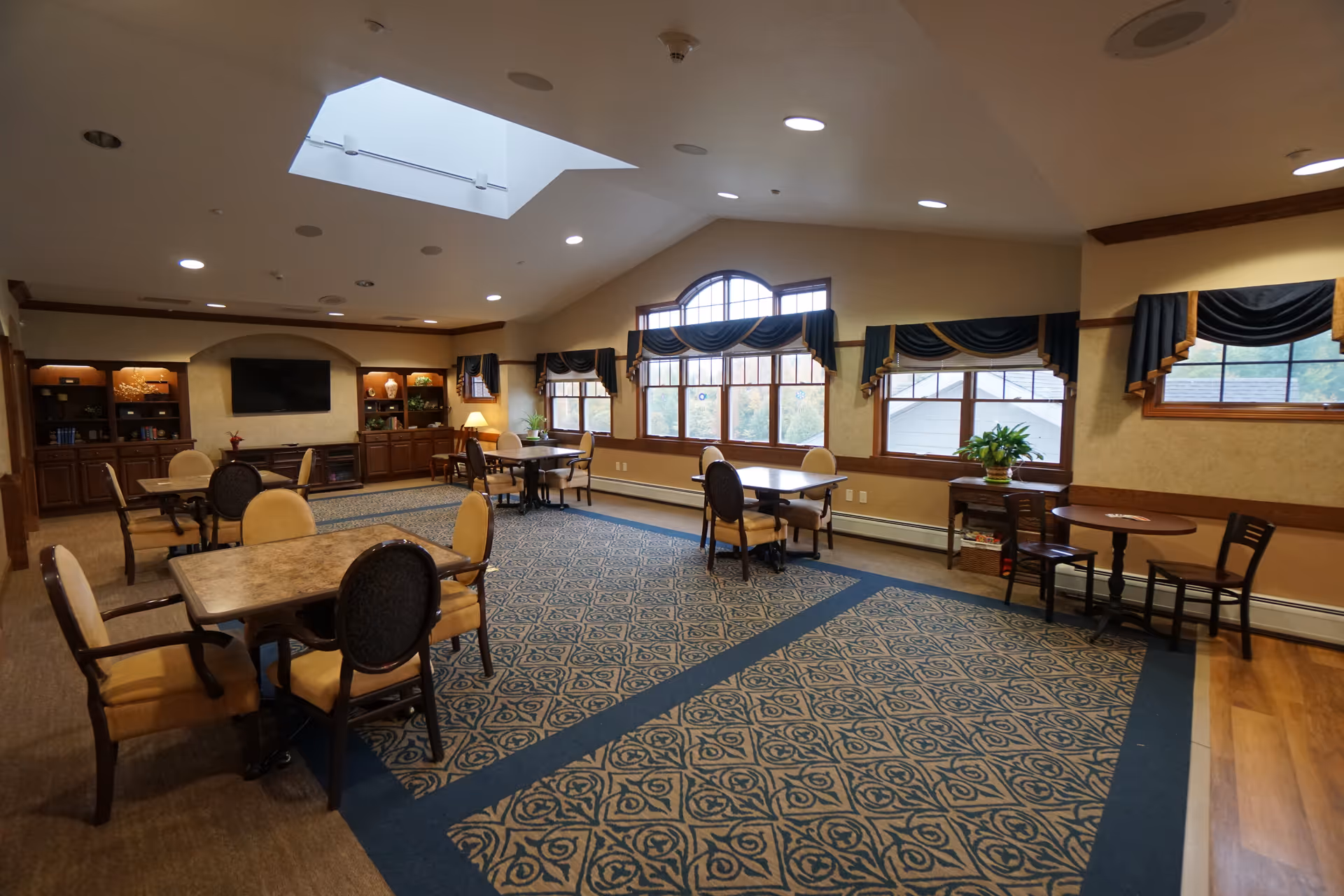 A spacious, well-lit common area with multiple tables and chairs arranged on a patterned carpet. Large windows with dark blue and gold valances allow natural light to fill the room. Built-in wooden cabinets and shelves line the far wall, with a flat-screen TV mounted above a fireplace. A skylight in the ceiling adds additional light to the room.