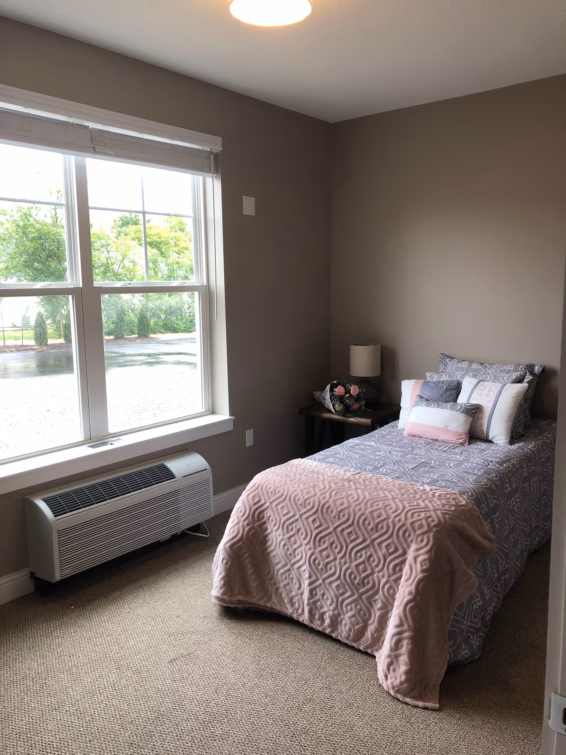 A small bedroom with a single bed covered in a gray and pink patterned bedspread and several pillows. Next to the bed is a small wooden nightstand with a lamp and a bouquet of flowers. A large window with white blinds lets in natural light, and there is a white air conditioning unit below the window. The walls are painted beige and the floor is carpeted.