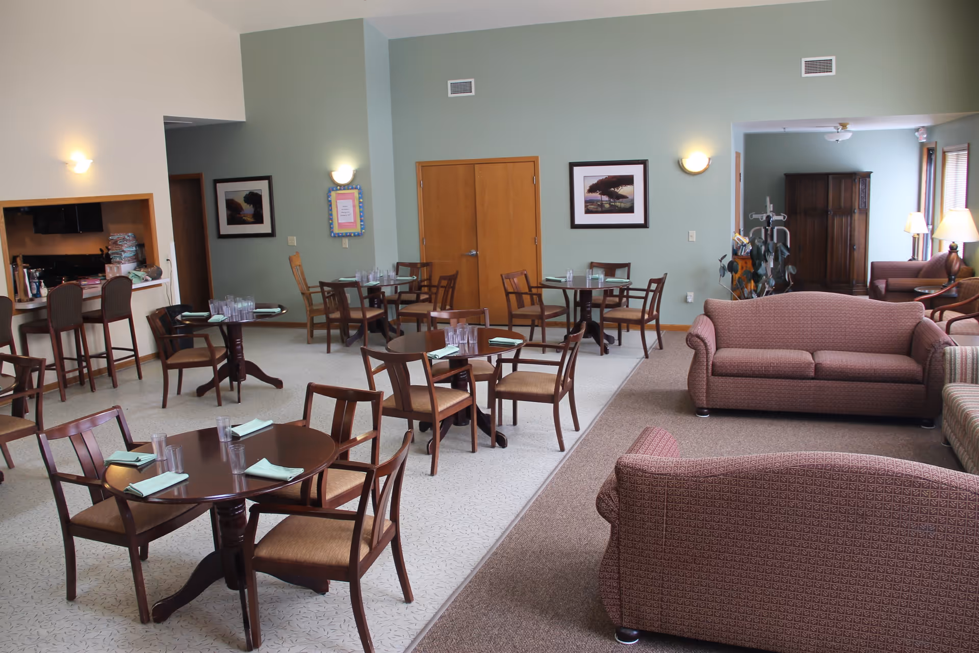 A spacious room with multiple round wooden tables and chairs set with glasses and napkins, adjacent to a seating area with patterned sofas and armchairs. The walls are painted light green and decorated with framed pictures. There is a kitchen pass-through window with bar stools on the left side and a wooden cabinet in the background near windows letting in natural light.