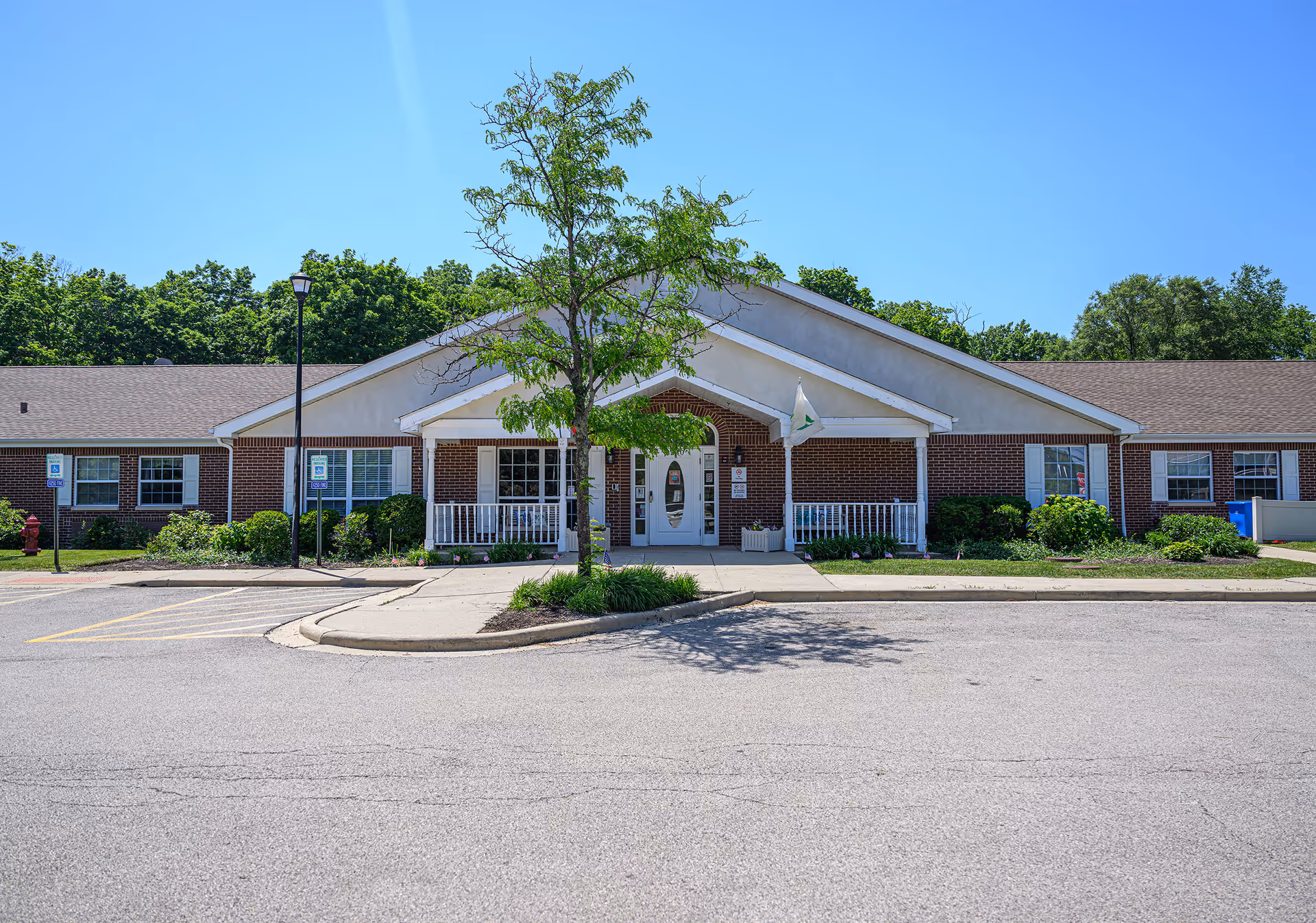 Front exterior view of a single-story brick building with a white entrance porch, a small tree in front, and a clear blue sky. The building has multiple windows and a parking area with handicap parking signs.