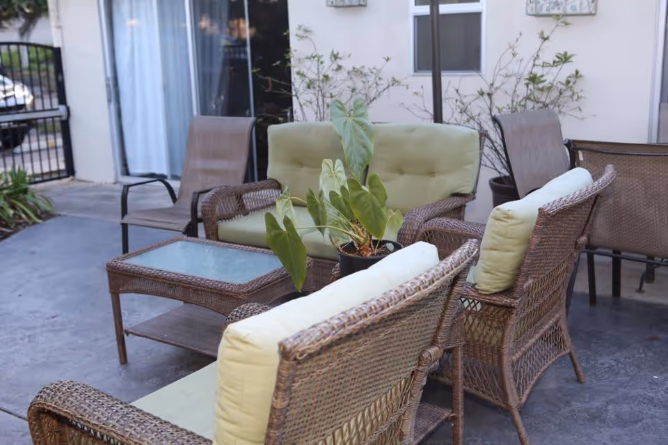 Outdoor patio area with wicker furniture including cushioned chairs and a loveseat, a glass-top coffee table, and a potted plant in the center. The patio is adjacent to a building with windows and a sliding glass door.
