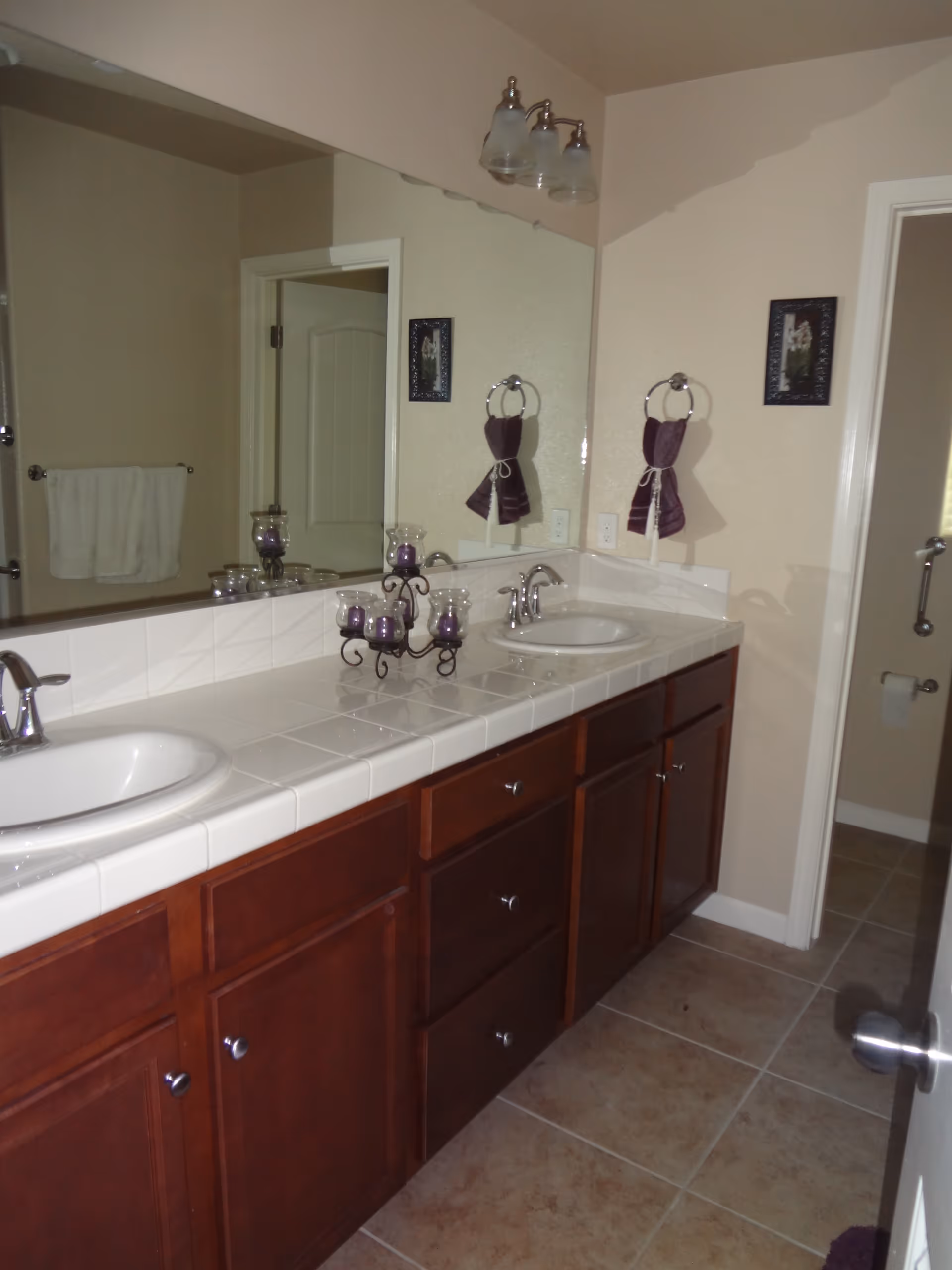 Bathroom with a double sink vanity featuring white tiled countertops and dark wooden cabinets. Above the sinks is a large mirror with three-light fixture. There are purple hand towels hanging on towel rings and a decorative candle holder with purple candles on the countertop. The floor is tiled and a door to a toilet area is visible.