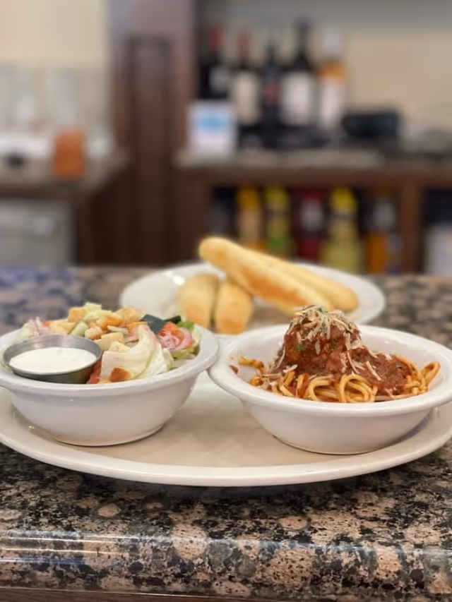 A plate with a bowl of salad with dressing, a bowl of spaghetti topped with a meatball and shredded cheese, and two breadsticks on a white plate on a granite countertop with a blurred background of bottles and kitchen items.