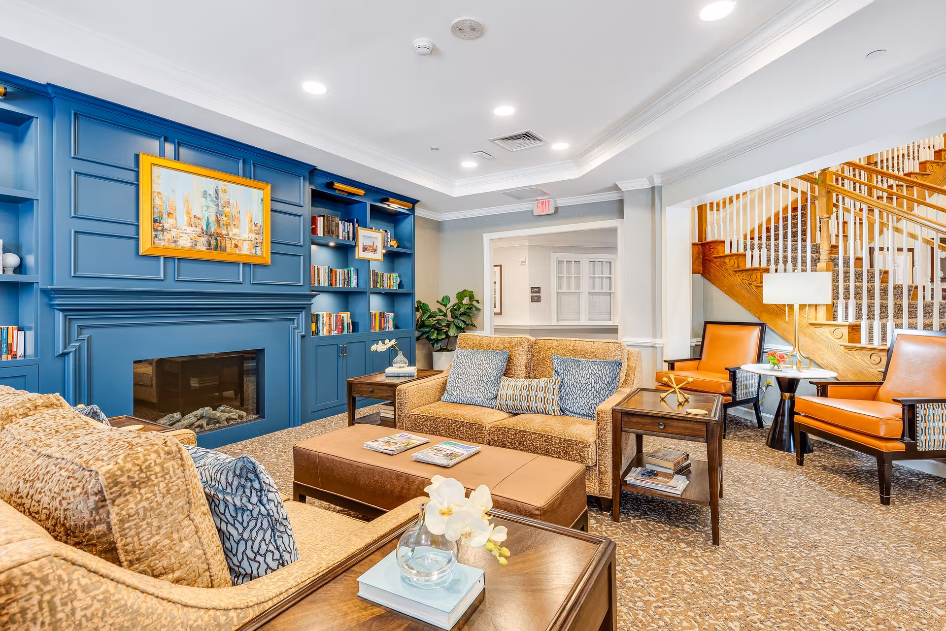 A cozy living room area with beige sofas adorned with patterned cushions, a brown ottoman coffee table, and wooden side tables. The room features a blue built-in bookshelf and fireplace with a colorful framed painting above it. In the background, there are two orange armchairs with a small round table between them, and a wooden staircase with white railings. The room is well-lit with recessed ceiling lights.