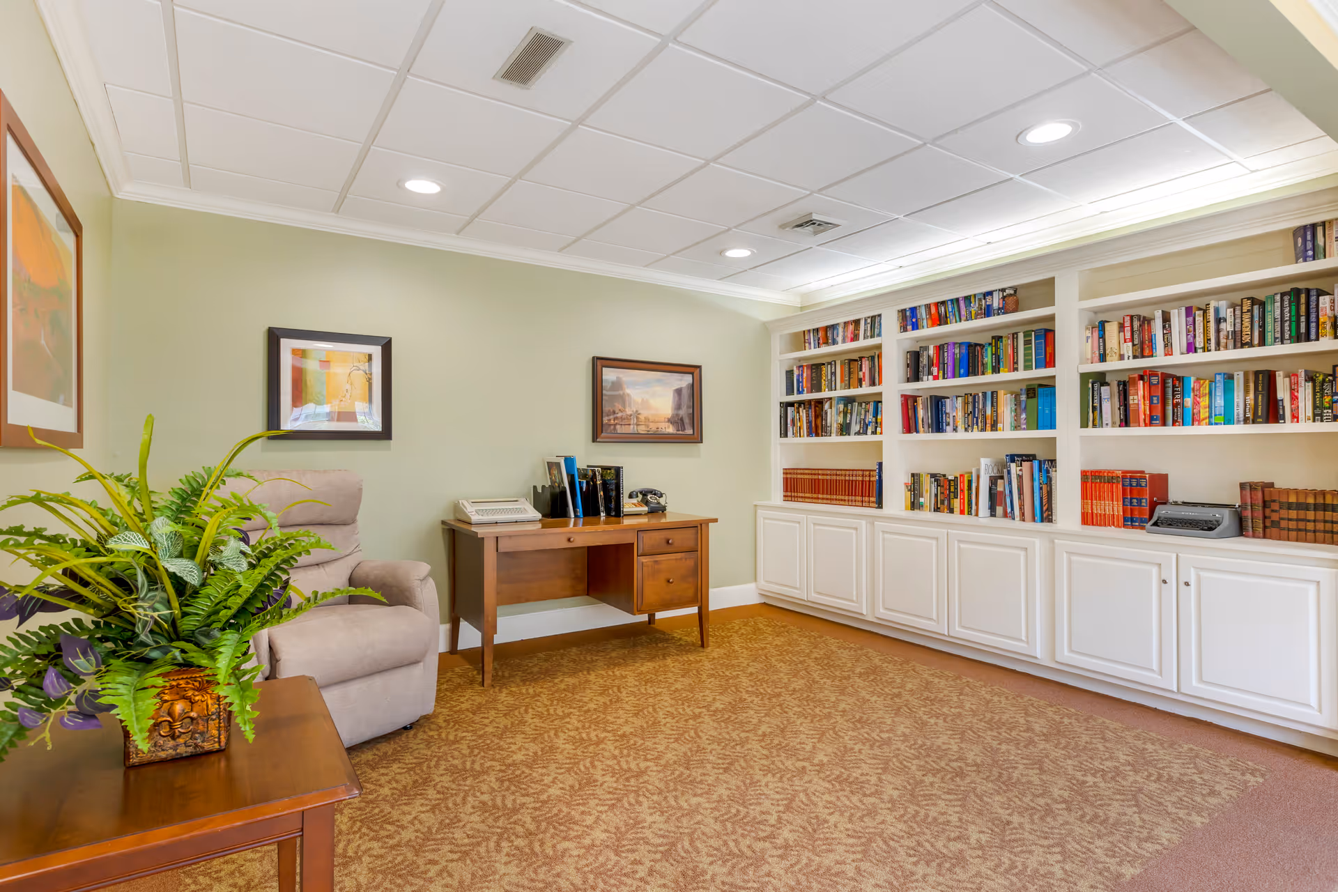 A cozy reading room with a beige recliner chair, a wooden desk with books and a vintage telephone, and built-in white bookshelves filled with books. There is a framed painting on the wall and a wooden side table with a potted green plant. The room has a patterned carpet and a white tiled ceiling with recessed lighting.