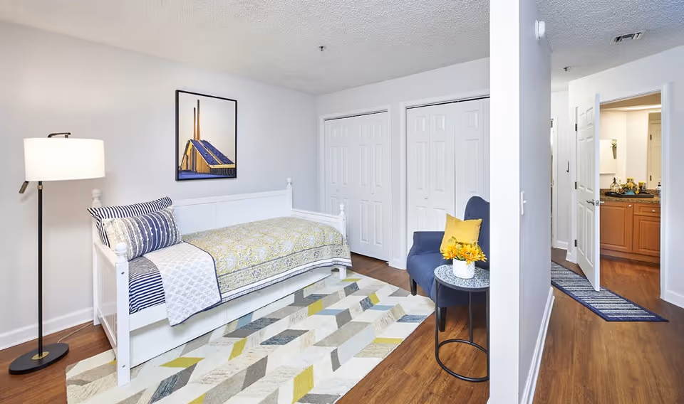 A bright bedroom with a white daybed covered with a patterned quilt and pillows, a floor lamp on the left, a framed picture on the wall above the bed, a geometric patterned rug on the wooden floor, a blue armchair with a yellow pillow and a small round table with a vase of yellow flowers. In the background, there are two white closets and an open door leading to a bathroom with wooden cabinets and a granite countertop.