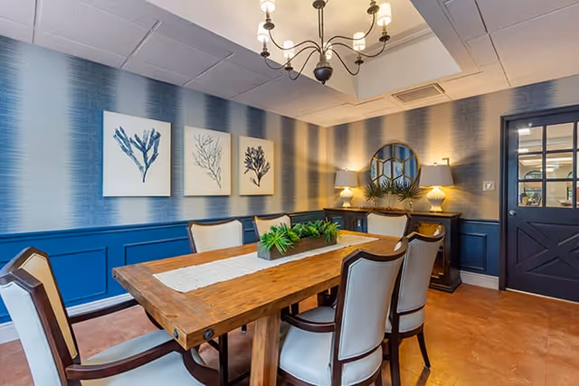 Well-lit dining room with a wooden table surrounded by upholstered chairs, blue-accented walls with coral artwork, and a chandelier.