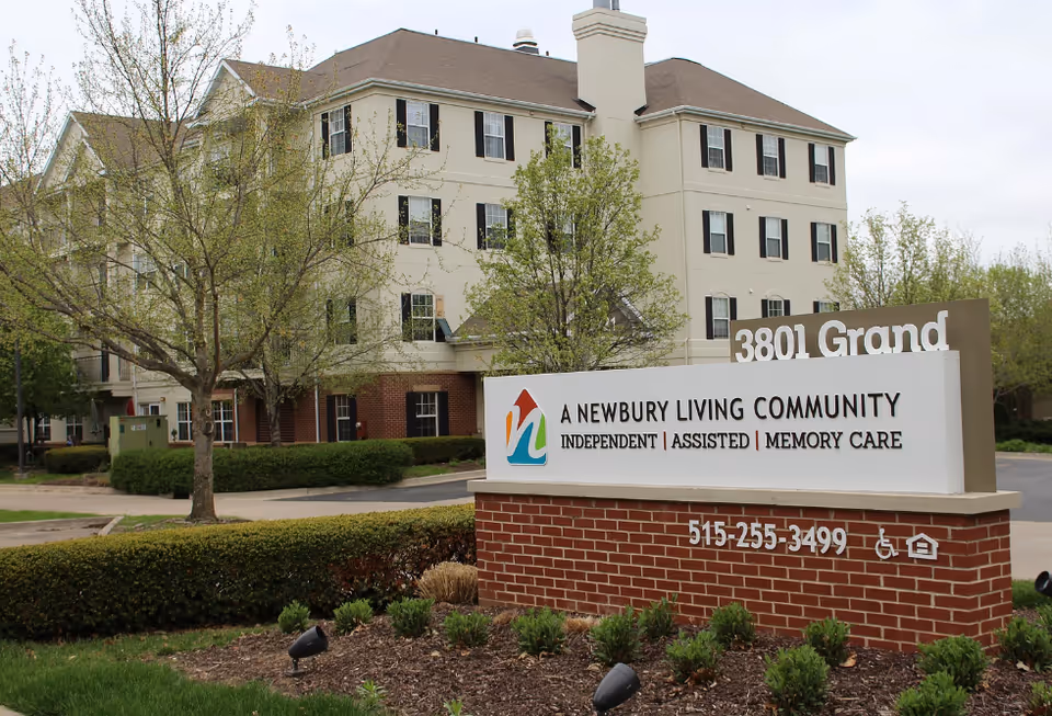 Exterior view of a multi-story beige building with black window shutters surrounded by trees and shrubs. In front of the building is a brick sign with a white panel displaying the logo and text: 'A Newbury Living Community Independent | Assisted | Memory Care' along with a phone number and accessibility symbols. The address '3801 Grand' is partially visible on a sign behind the main sign.