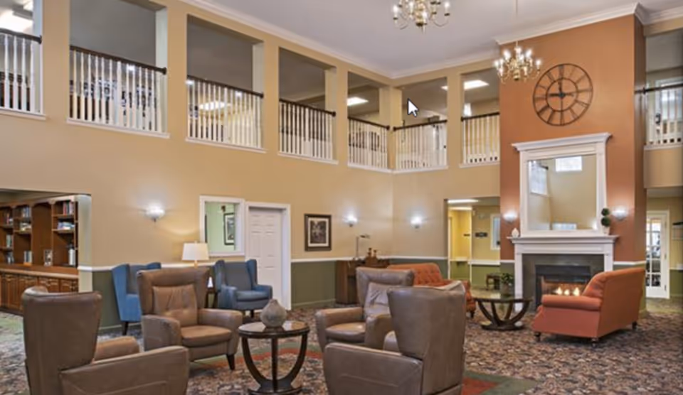 A spacious senior living facility common area with multiple comfortable armchairs arranged around small round tables. The room features a high ceiling with a second-floor balcony railing overlooking the area. There is a large wall clock above a fireplace with a mirror above the mantel. The walls are painted in warm tones, and the room is well-lit with wall sconces and chandeliers.