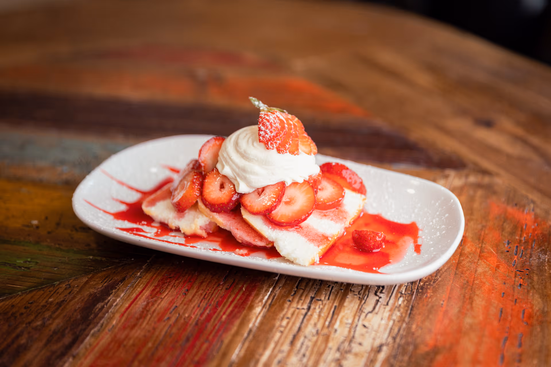A dessert plate with slices of cake topped with sliced strawberries, whipped cream, and strawberry sauce on a rustic wooden table.