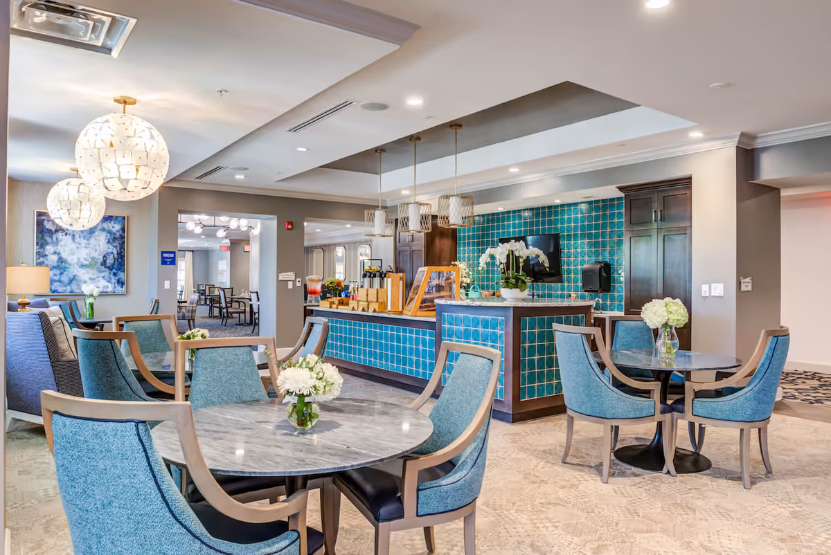 Bright dining/lounge area with round tables, blue upholstered chairs, and a tiled serving counter with pendant lights.
