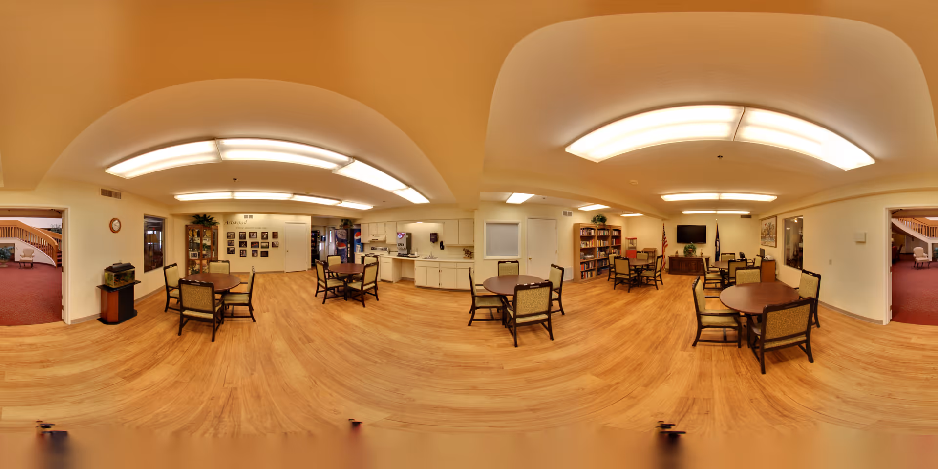 A spacious, well-lit common area with wooden flooring and multiple round tables surrounded by chairs. The room features a bookshelf, a television mounted on the wall, and an American flag. There is a small kitchenette area with cabinets and a countertop. The walls are decorated with framed pictures and a clock. Two doorways lead to adjacent rooms with red carpeting and seating areas.