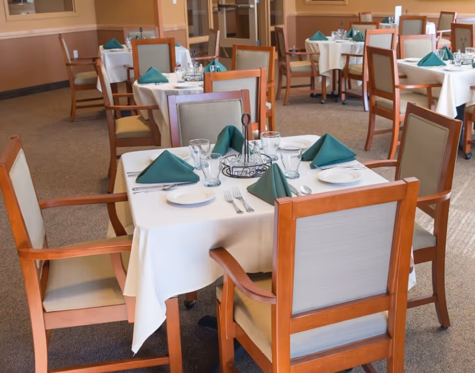 A dining room with several wooden tables and chairs. Each table is covered with a white tablecloth and set with green folded napkins, plates, glasses, and silverware. The room has beige walls and carpeted floors.