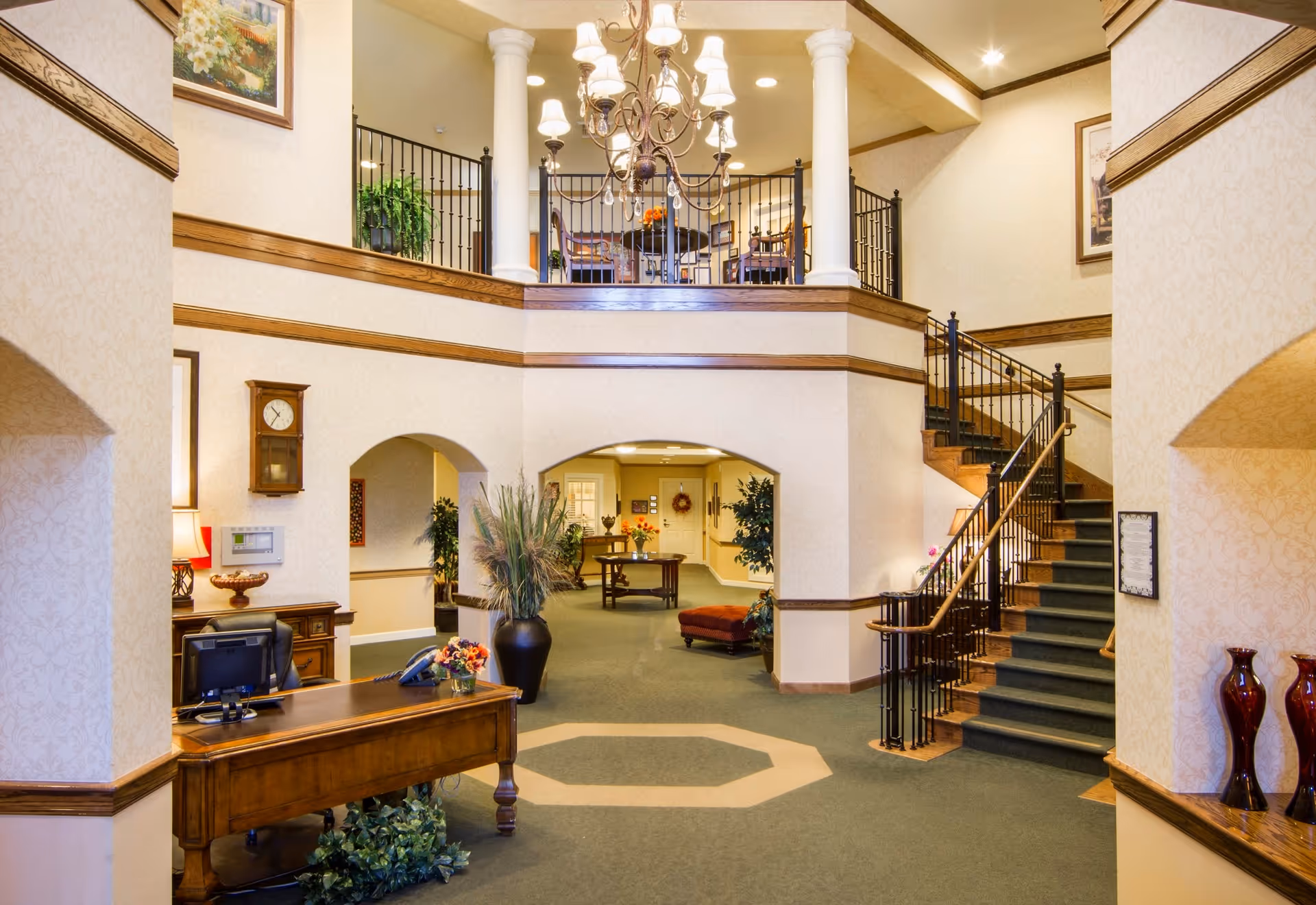 Interior view of a senior living facility lobby with a wooden reception desk, a computer, decorative plants, a staircase with black railings leading to an upper floor, and a chandelier hanging from the ceiling. The walls are light-colored with wooden trim, and there are framed pictures and a wall clock visible.
