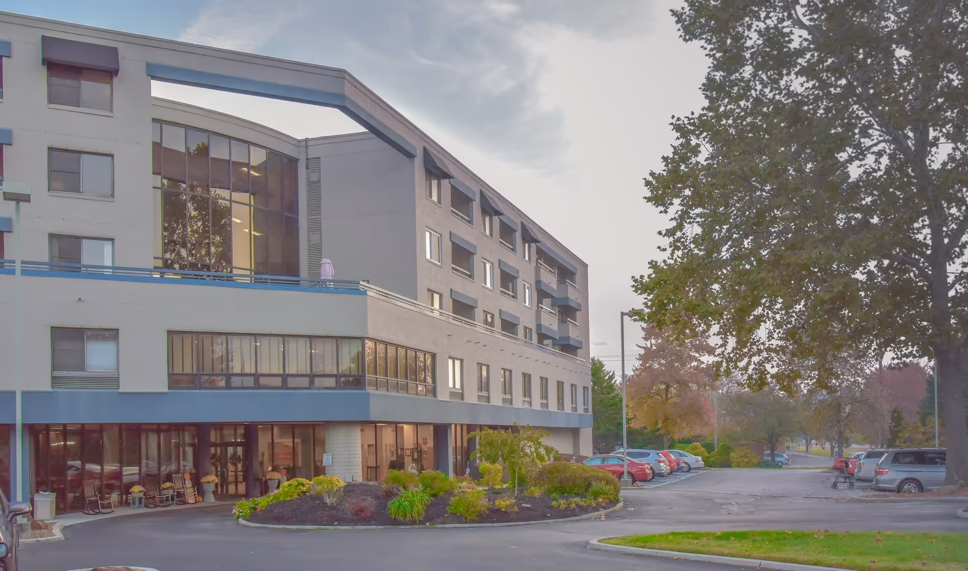 Exterior view of a multi-story senior living facility building with large windows and a landscaped area with shrubs and trees in front. Several cars are parked in the parking lot adjacent to the building under a cloudy sky.