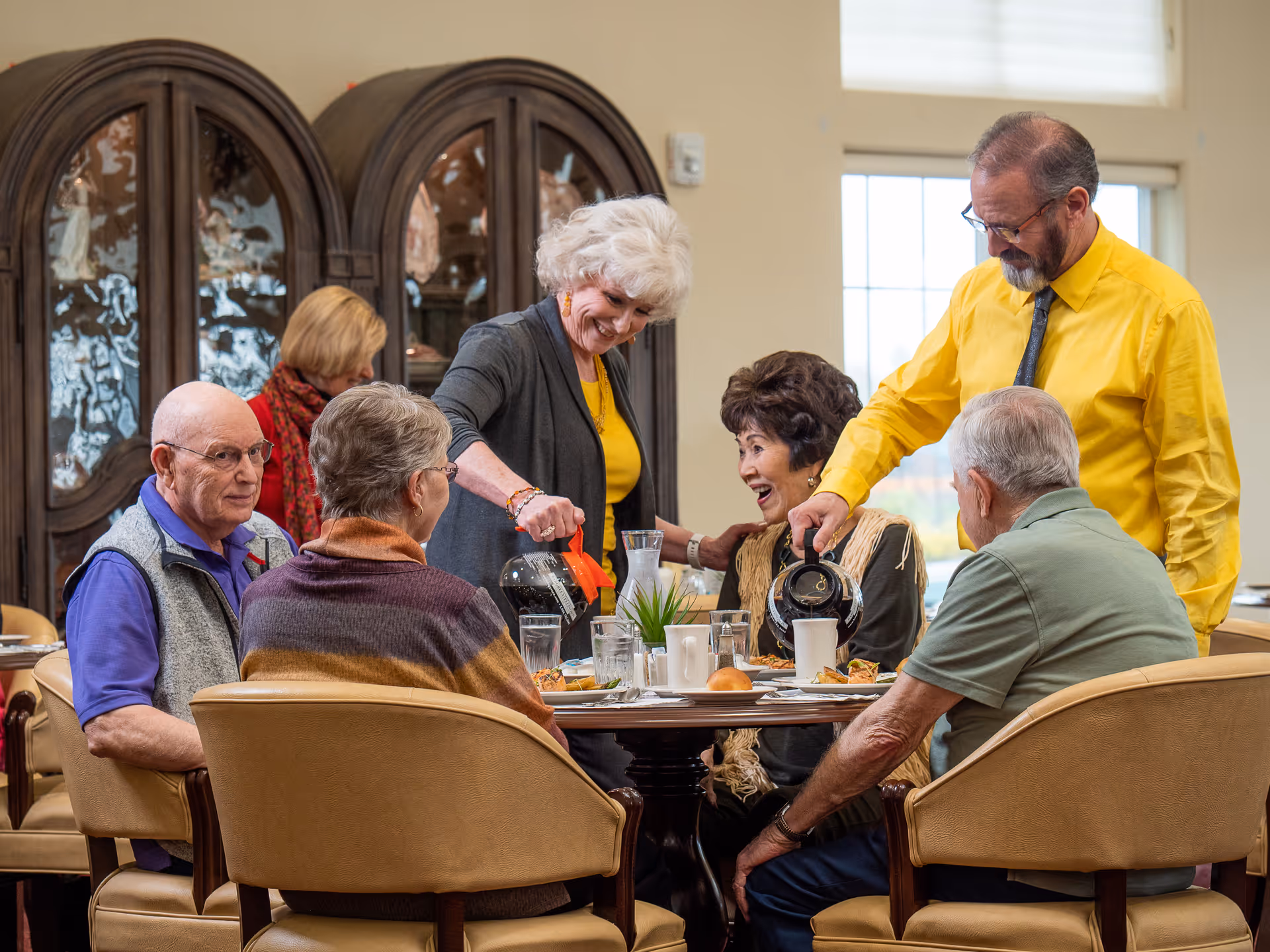 A group of six elderly people sitting around a dining table in a senior living community. Two of them are pouring coffee while others are engaged in conversation and smiling. The room has large windows and a wooden cabinet with glass doors in the background.