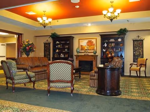 A warm, furnished common room with sofas and armchairs arranged around a fireplace and decorative shelving under chandeliers.