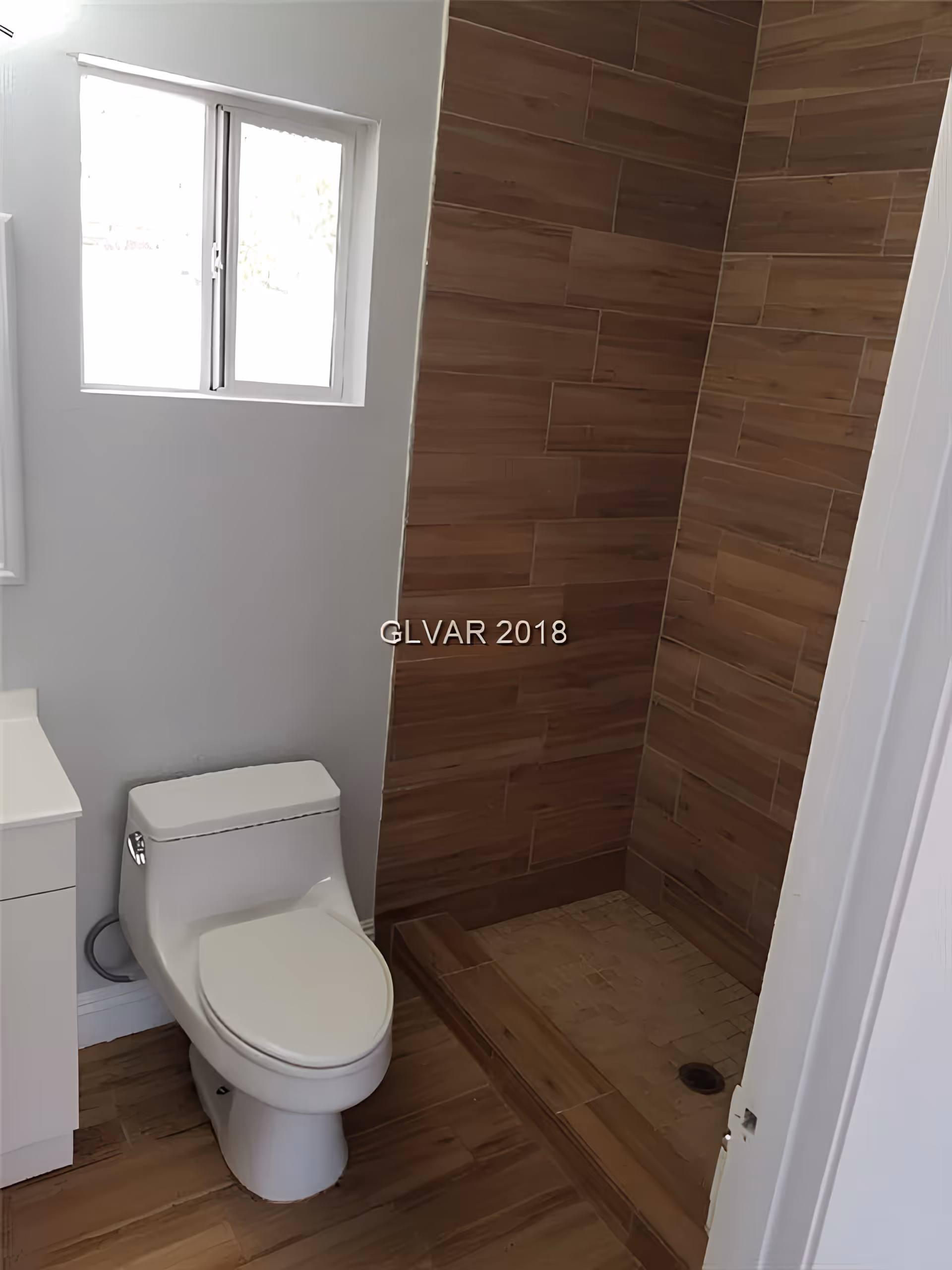 A bathroom with a white toilet next to a wooden-tiled walk-in shower. There is a small window above the toilet and a white vanity partially visible on the left side.